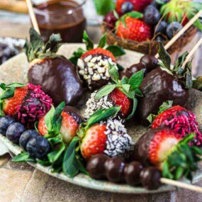 A plate with chocolate-covered strawberries, skewered berries, and various garnished fruit berries on a cloth napkin. A bowl with more berries and chocolate sauce in the background.