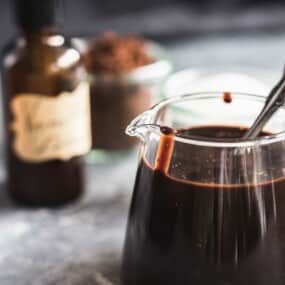 A glass pitcher filled with a dark liquid has a spoon inside. In the background, there are small jars containing powder and a brown bottle with an illegible label.