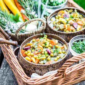 A wicker basket with three bowls of mixed vegetables and herbs, alongside fresh carrots and greens, sits on a wooden surface outdoors.