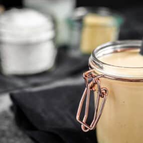 A jar filled with creamy sauce or dressing is placed on a dark cloth with a black spoon inside the jar. In the background, there are blurry containers holding various ingredients.
