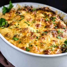A white ceramic dish filled with a baked casserole topped with melted cheese and garnished with fresh parsley.
