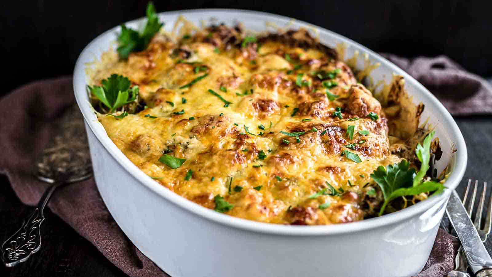 A white ceramic dish filled with a baked casserole topped with melted cheese and garnished with fresh parsley.