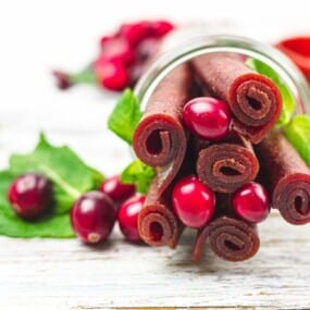 Rolled fruit leather sticks and fresh cranberries on a white wooden surface with green leaves, some cranberries, and a red cloth in the background.
