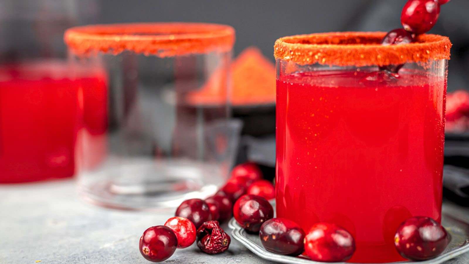 Two glasses with red juice, garnished with cranberries and an orange sugar rim, sit on a gray surface with scattered cranberries.