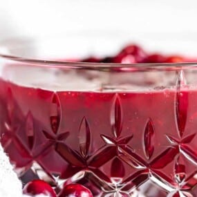 A clear, intricately patterned glass bowl filled with dark red cranberry sauce, garnished with whole cranberries.