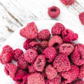 A bowl of dried raspberries on a wooden surface, with several dried raspberries scattered around the bowl.