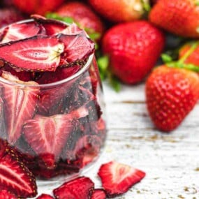 A glass filled with dried strawberries is placed on a wooden surface, surrounded by fresh strawberries.