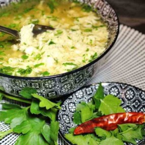 A hand holds a spoon over a bowl of soup garnished with herbs, placed on a patterned placemat. A smaller bowl with a red chili pepper and more greens is beside it.