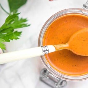 An open jar of orange-red sauce with a spoon resting on top. Parsley leaves are visible to the side on a white surface.