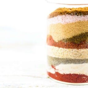 A glass jar filled with various layers of colorful spices arranged in stripes, placed on a light-colored surface with a white background.