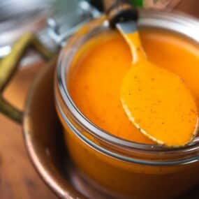 A spoon rests on a jar filled with bright orange sauce placed on a wooden surface.