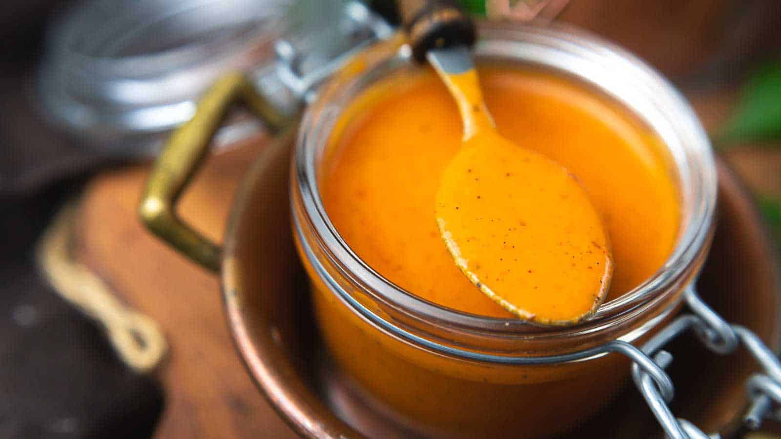 A spoon rests on a jar filled with bright orange sauce placed on a wooden surface.