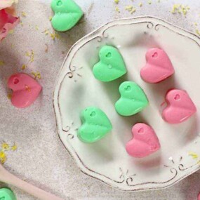 A white plate contains pastel green and pink heart-shaped candies. A pink rose is in the upper left corner, and a wooden spoon with yellow zest is at the bottom left. More candies are scattered around.