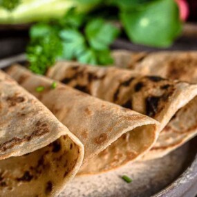 A plate with four folded, slightly charred flatbreads garnished with chopped herbs. In the background, leafy greens and vegetables are faintly visible.