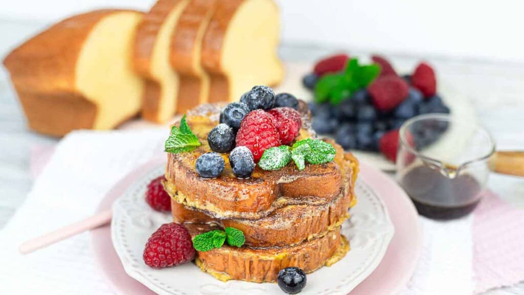A dish of French toast topped with powdered sugar, mint leaves, raspberries, blueberries, and blackberries; maple syrup and sliced bread are in the background.