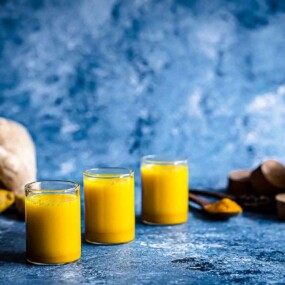 Three glasses of yellow juice are in the foreground with fresh ingredients and a pitcher of juice in the background, all set against a blue textured surface.