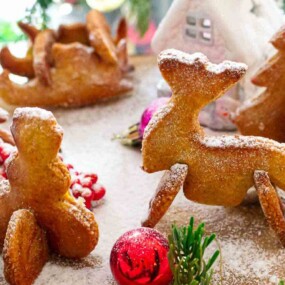 Festive gingerbread cookies shaped like reindeer and trees, dusted with powdered sugar, surrounded by colorful ornaments and greenery.