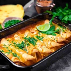 A black baking dish filled with enchiladas topped with herbs and a lime wedge. Surrounding the dish are avocado slices, tortillas, lime halves, a jar of sauce, and fresh parsley.