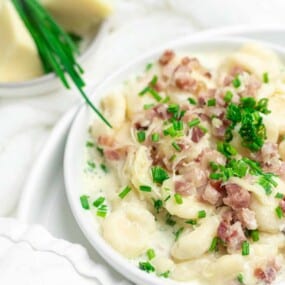 A bowl of creamy gnocchi topped with chopped chives and diced bacon sits on a white plate with a fork. In the background, sliced cheese and chives are on a small plate.