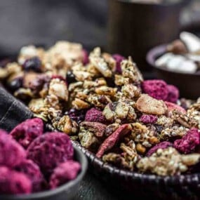 A wooden tray of mixed granola with dried berries is displayed on a dark surface, with bowls of similar snacks in the background.