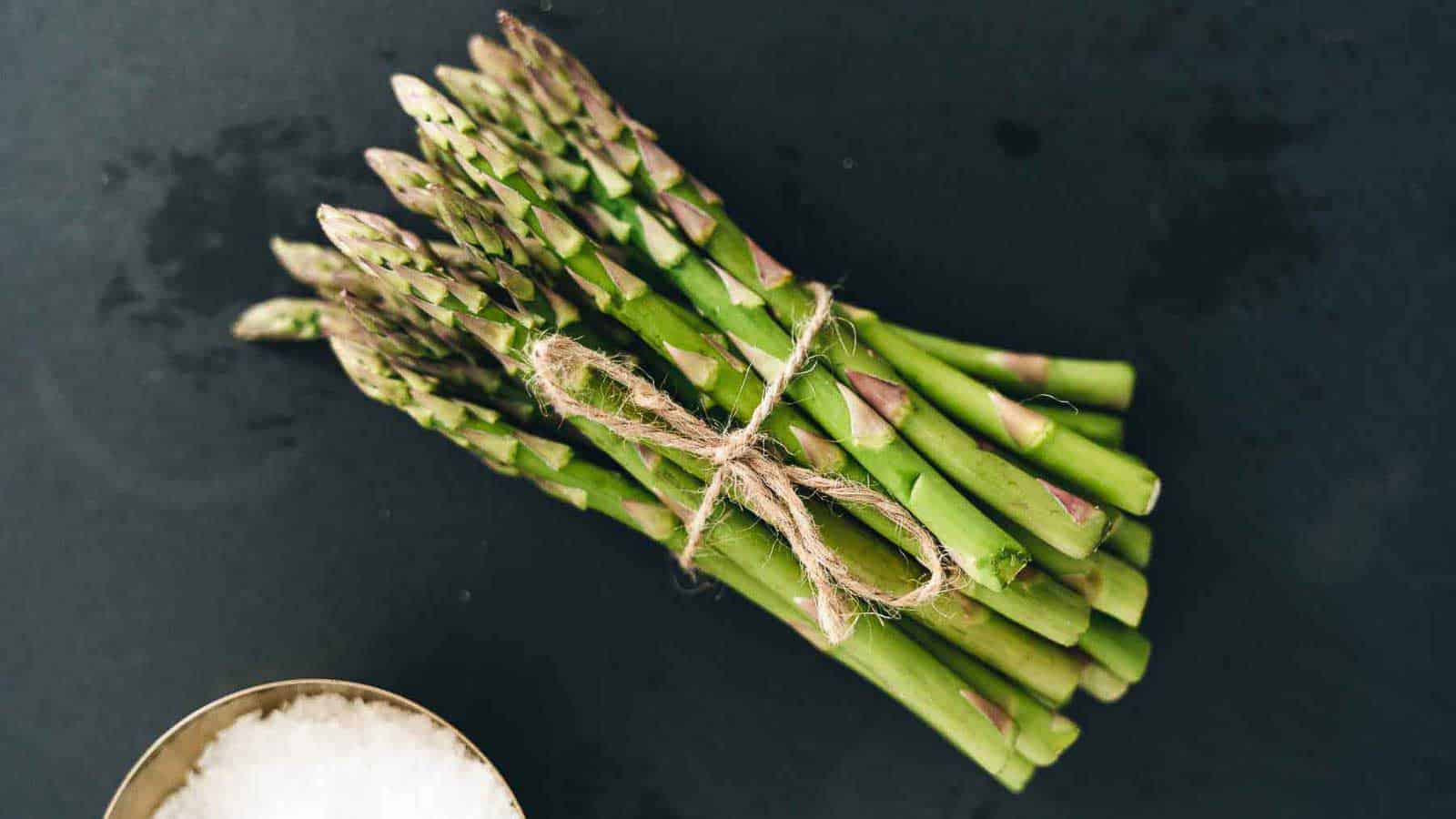 A bunch of green asparagus stalks tied with twine, placed on a dark surface. A small bowl of coarse salt is partially visible in the bottom left corner.