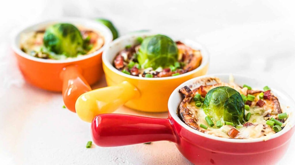 Three small cups with handles, each containing a baked dish topped with a Brussels sprout, chopped green onions, and bits of bacon, placed in a row on a white surface.