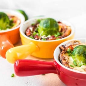 Three small cups with handles, each containing a baked dish topped with a Brussels sprout, chopped green onions, and bits of bacon, placed in a row on a white surface.