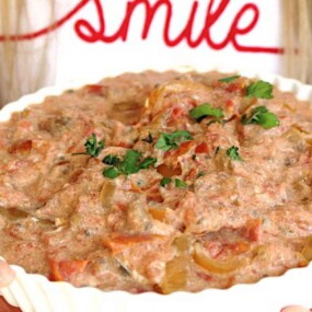 A person holds a dish of creamy mixed food topped with cilantro. The person wears a white shirt with the word "smile" in red cursive.