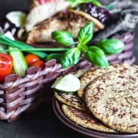 A basket of fresh vegetables including tomatoes, cucumbers, and lettuce sits beside a plate with three pieces of flatbread. Fresh basil leaves garnish the basket.