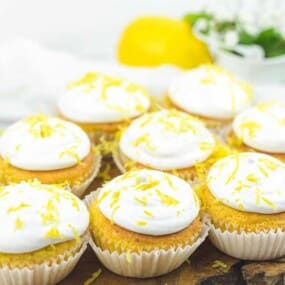 Nine lemon cupcakes with white frosting and lemon zest on top are arranged on a wooden board. A lemon and small white flowers are in the background.