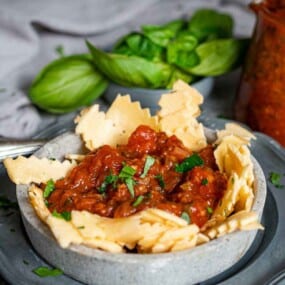 A bowl of pasta topped with tomato sauce and garnished with fresh herbs. A jar of tomato sauce and basil leaves are visible in the background.