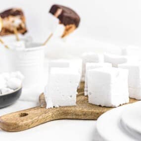 A wooden board with large marshmallows, a black bowl with smaller marshmallows, and a cup of hot chocolate. Marshmallows coated with chocolate on sticks are in the background.