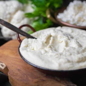 A bowl of cauliflower mash with a spoon, placed on a wooden board. In the background, there are pieces of raw cauliflower and a bowl of crumbled cauliflower.