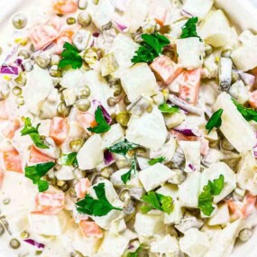 A bowl of mixed vegetable salad with potatoes, carrots, peas, and parsley, dressed in a creamy sauce.