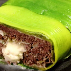 A close-up of a meatloaf wrapped in bright green leek leaves, with a visible pocket of melted cheese in the center.