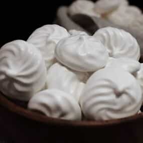 A bowl filled with white meringue cookies, displaying their smooth, airy texture against a dark background.