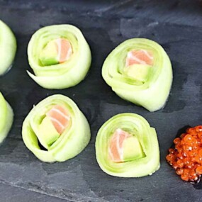 A platter of sushi rolls made with cucumber wraps, containing salmon and avocado, accompanied by wasabi, ginger, and fish roe on a slate surface.