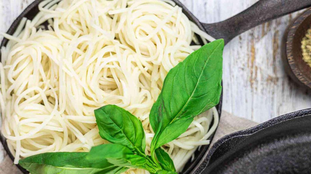 A cast iron skillet filled with cooked spaghetti and garnished with fresh basil leaves. A partial view of another skillet and a bowl of grated cheese are in the background.