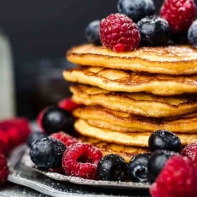 A stack of pancakes topped with fresh raspberries and blueberries on a plate, surrounded by more berries, with a small jug and a bottle in the background.