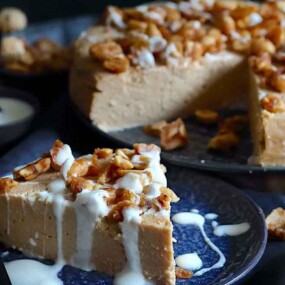 A slice of peanut-topped cheesecake drizzled with white sauce on a dark plate, with a whole cheesecake and more peanuts in the background.