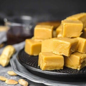 A stack of caramel fudge pieces is arranged on a black plate with scattered nuts and a glass container in the background.