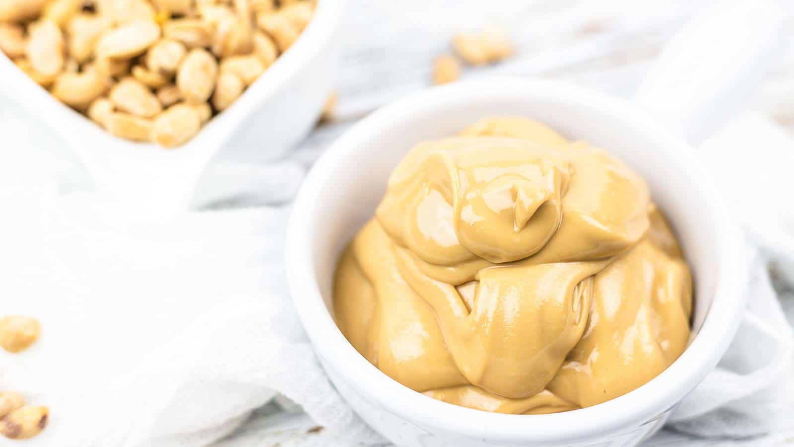 A white bowl filled with creamy peanut butter is placed next to a bowl of shelled peanuts.