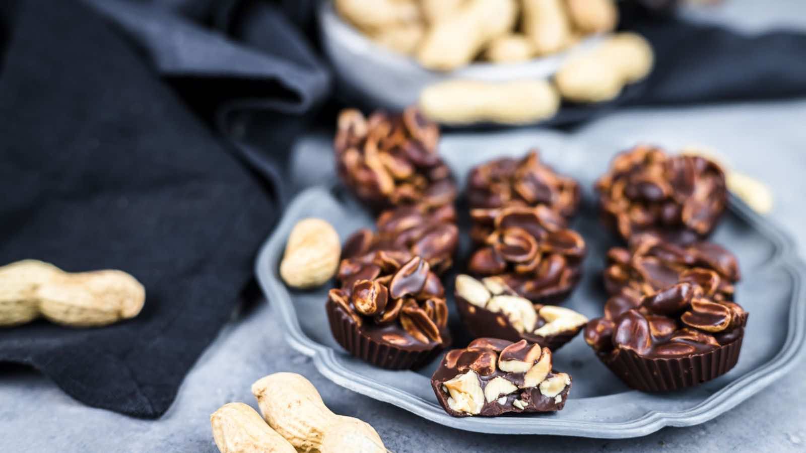 Chocolate peanut clusters on a plate with whole peanuts scattered around on a gray surface.