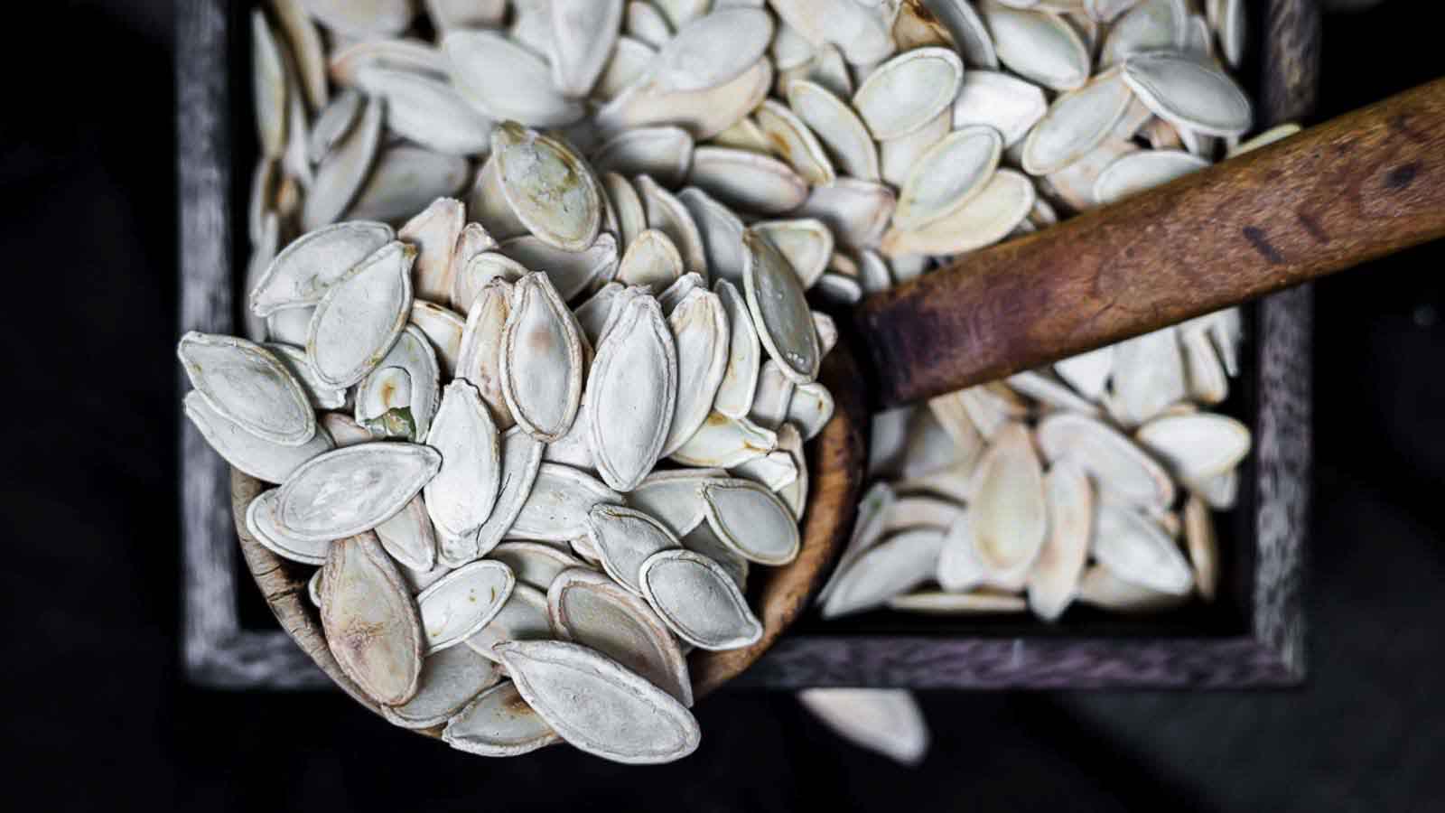 A close-up of a scoop filled with pumpkin seeds, with additional seeds filling a wooden container in the background.