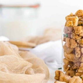 A clear glass filled with caramel-coated popcorn pieces is placed on a light-colored cloth, with a blurred jar and white background.