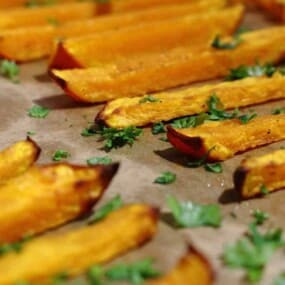 Close-up of roasted butternut squash fries topped with parsley, arranged on a baking sheet lined with parchment paper.