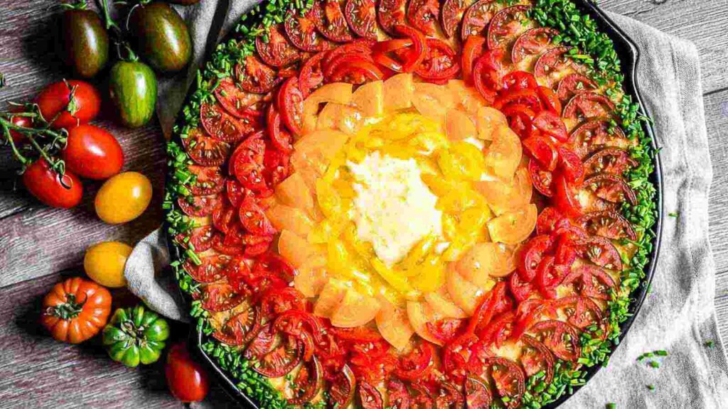 A vibrant dish featuring concentric rings of sliced tomatoes in red, orange, and yellow, topped with herbs and centered around a dollop of cream or cheese, placed on a wooden surface.