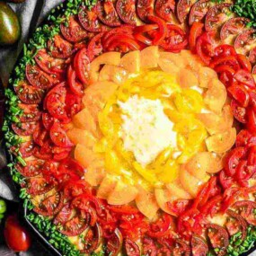 A vibrant dish featuring concentric rings of sliced tomatoes in red, orange, and yellow, topped with herbs and centered around a dollop of cream or cheese, placed on a wooden surface.