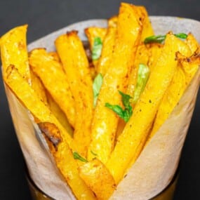A paper cone filled with seasoned yellow vegetable sticks, likely made from squash or a similar vegetable, garnished with green herbs.