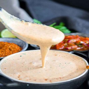 A spoon drizzles creamy sauce into a bowl, surrounded by various condiments including a spice mix, green herbs, and a chunky salsa in small bowls.